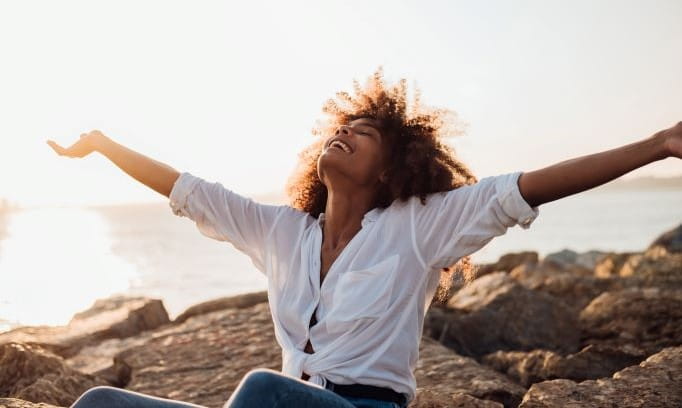 woman with arms thrown wide while sitting on rocks near water